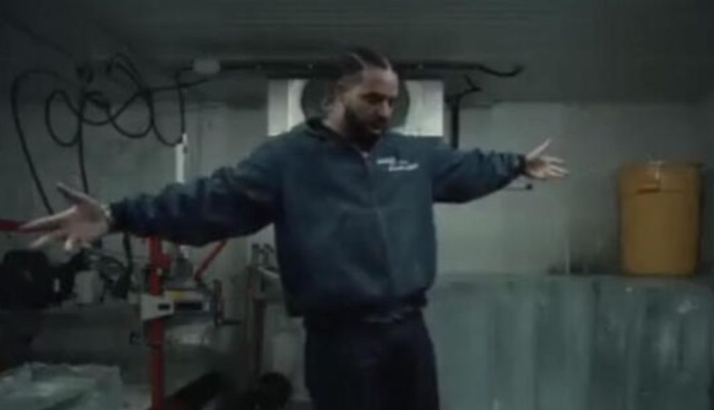Drake standing with arms outstretched inside an industrial-looking room, wearing a dark jacket and braided hair, looking down